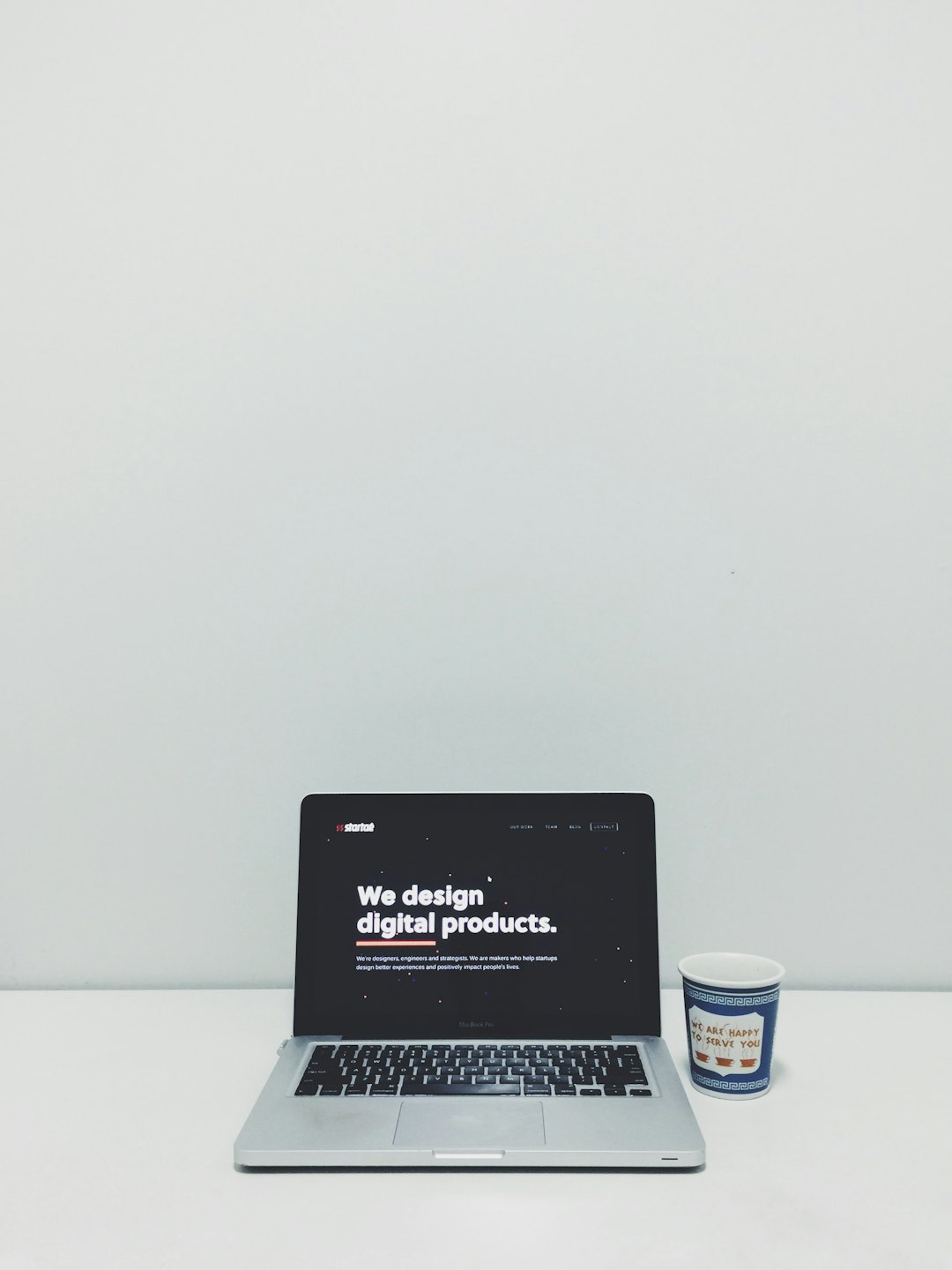 Laptop perak menampilkan 'Kami merancang produk digital' di atas meja putih.